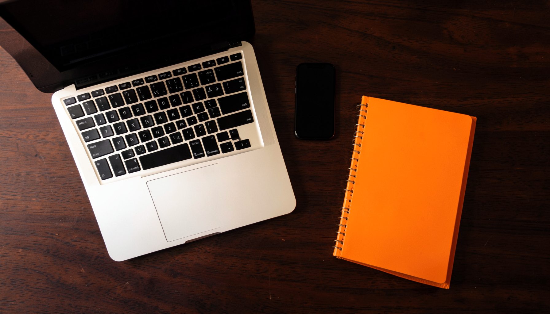 Overhead flat-lay photograph of a laptop, smartphone, and a notebook arranged on a dark wooden desk, with warm ambient light and subtle orange accents.