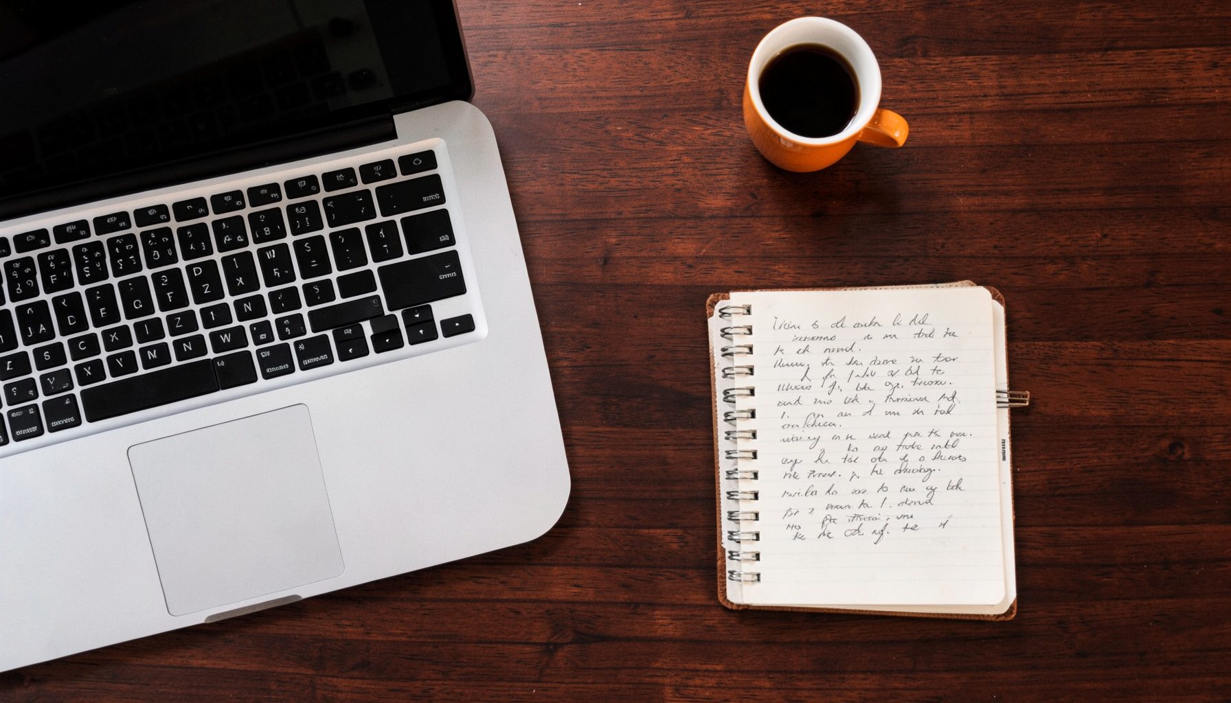 Overhead flat-lay photograph of a laptop, smartphone, and a handwritten notebook on a warm dark wooden desk, with a cup of coffee nearby; the scene feels calm, intentional, and ready for action, with deep browns and soft ambient light.