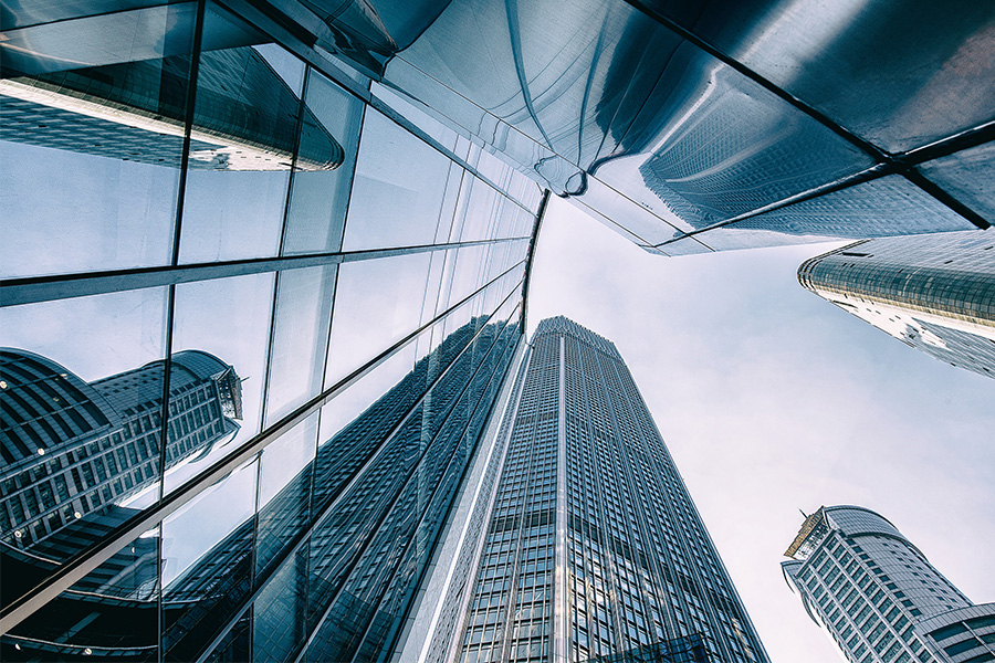 Weitwinkel-Aufnahme von unten, die eine Gruppe moderner Glas- und Stahlhochhäuser zeigt, deren reflektierende blaue und silberne Fassaden sich in einem hellen Himmel verlieren.